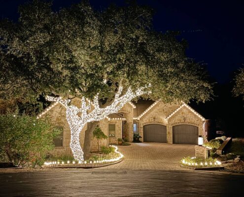 Front yard oak tree wrapped in white lights with lit landscape beds and roofline