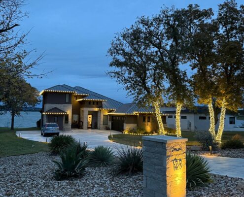 Lakefront home at dusk with trees wrapped in Christmas lights and a lit address pillar
