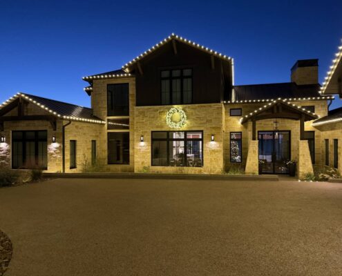 Warm white roofline Christmas lights and a lit wreath on a stone and timber home