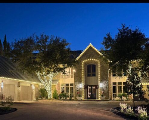 Stone estate with gables and arched entry outlined in Christmas lights and lit trees