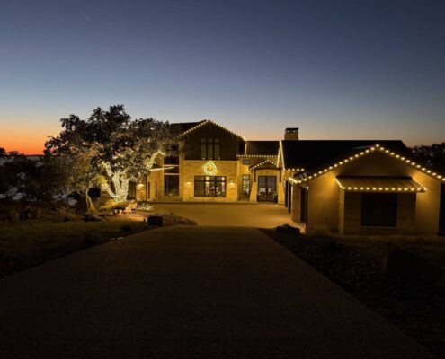Stone Hill Country home with roofline Christmas lights, wreath and lit oak tree at sunset