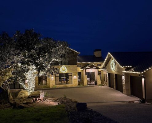 Stone home with roofline Christmas lights, lit wreaths and a wrapped oak tree at night