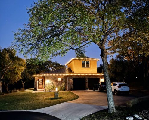 Two-story home with warm white roofline lights at dusk beneath a large oak